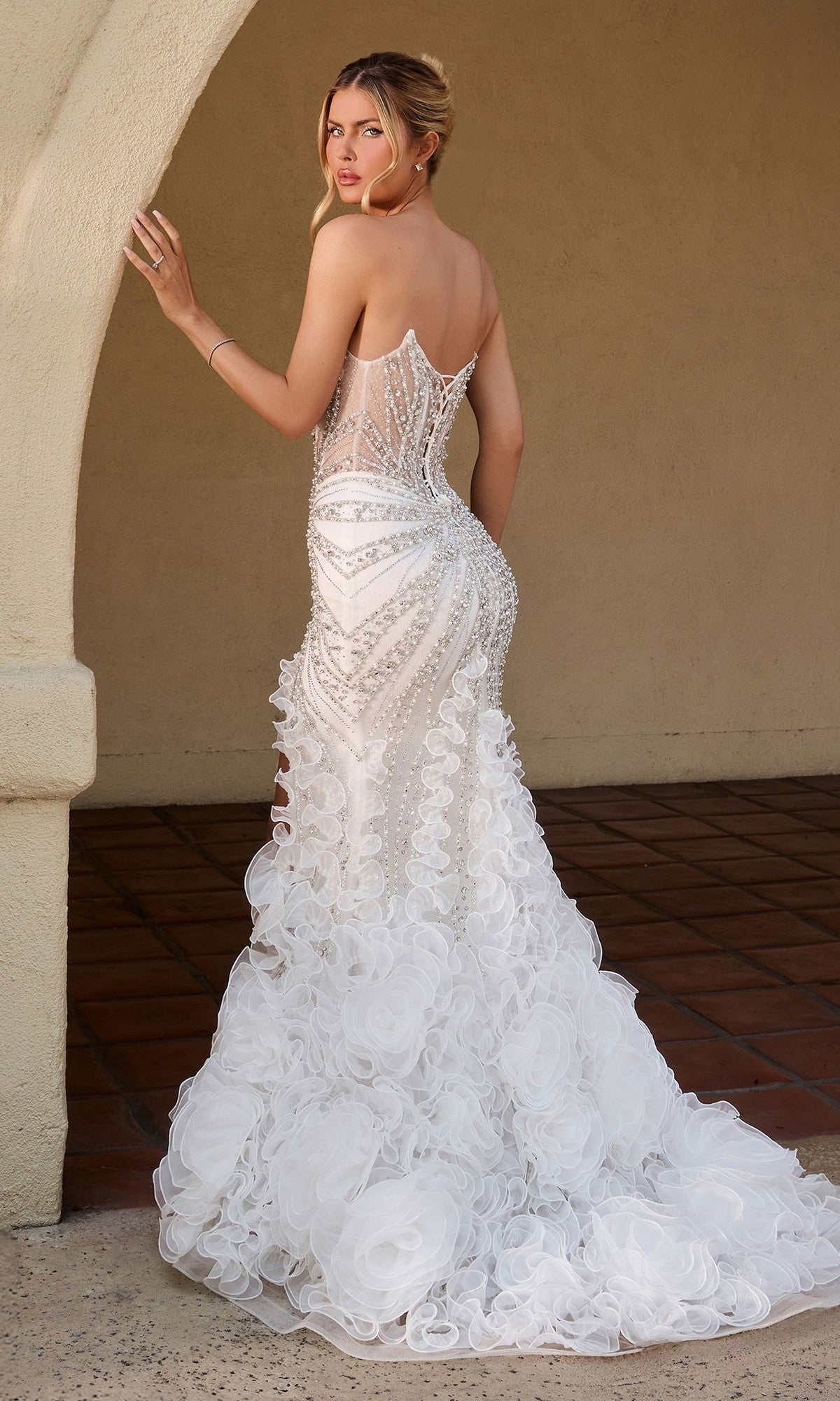 A woman stands by an archway, facing away from the camera, wearing the Ladivine CC2488W long formal dress—a strapless mermaid gown with sheer panels, bead embellishments, and a textured ruffled train.